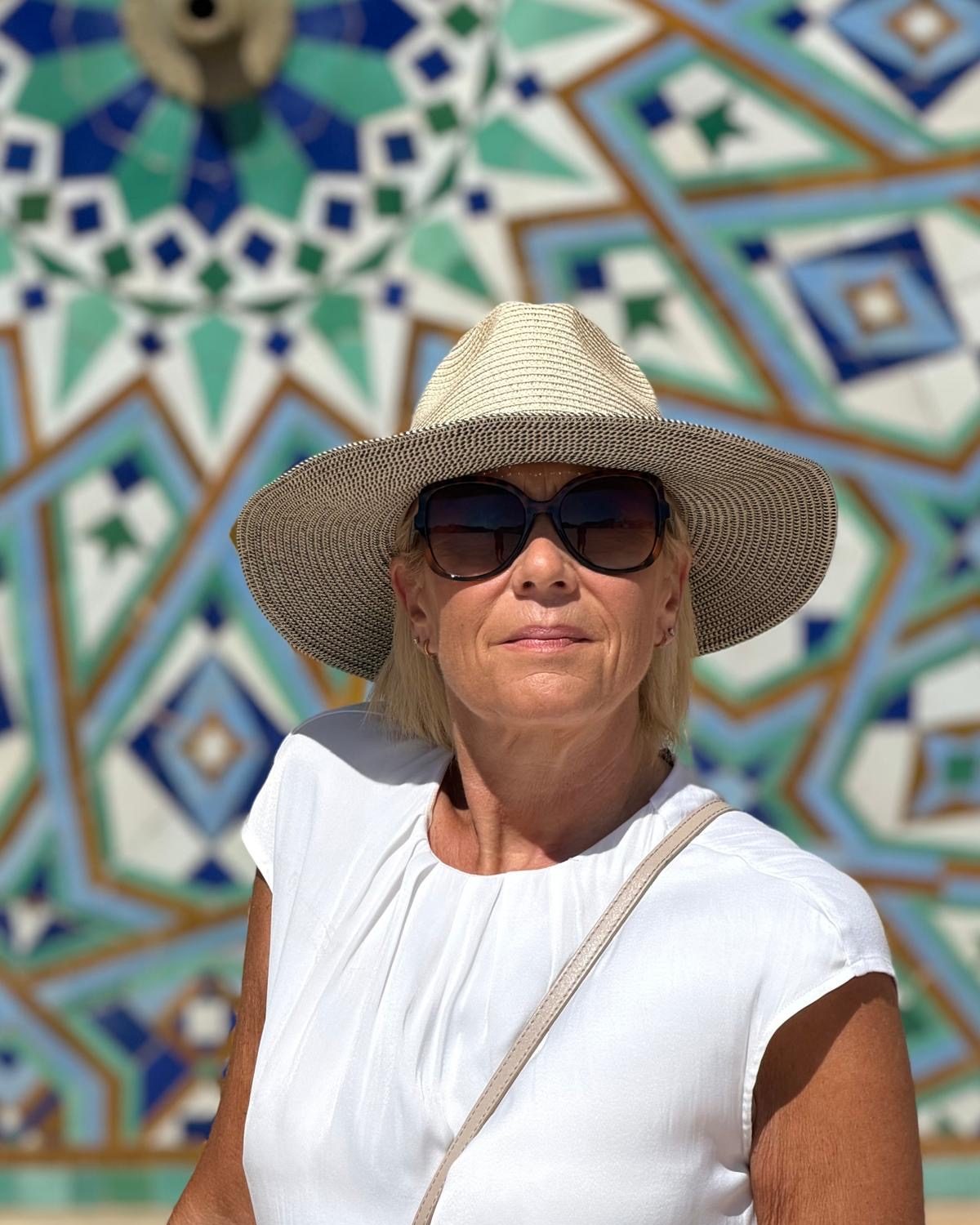 Nik in a natural hat with a speckled brim, sunglasses and a white top, in front of a blue and green mosaic tiled wall