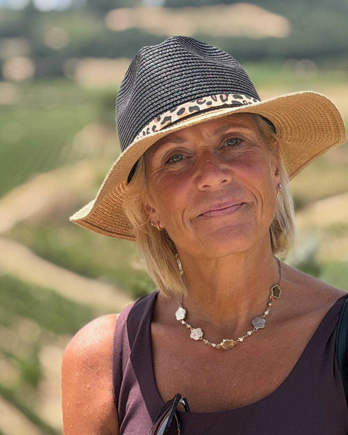 Nik smiling in a black and natural two-tone hat with a leopard-print band, with vineyards behind her