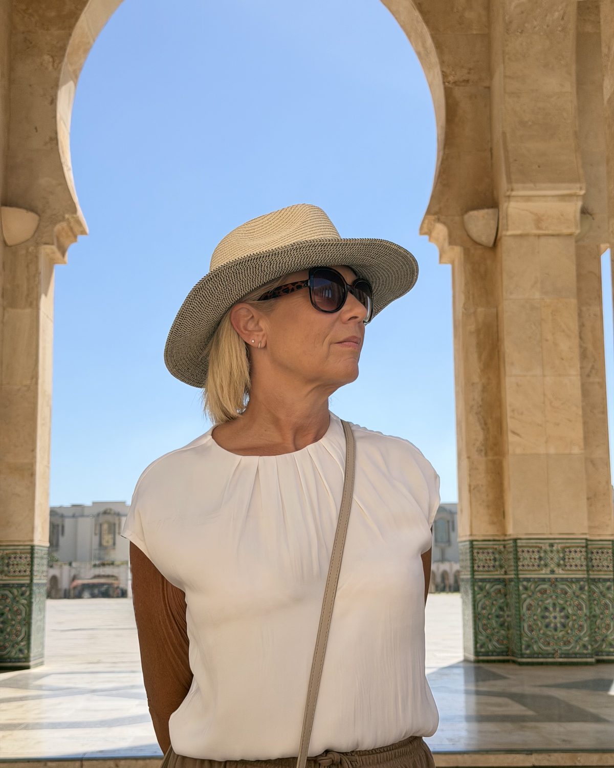 Nik in a natural speckled-brim hat and sunglasses, looking to the side beneath a stone archway against a blue sky